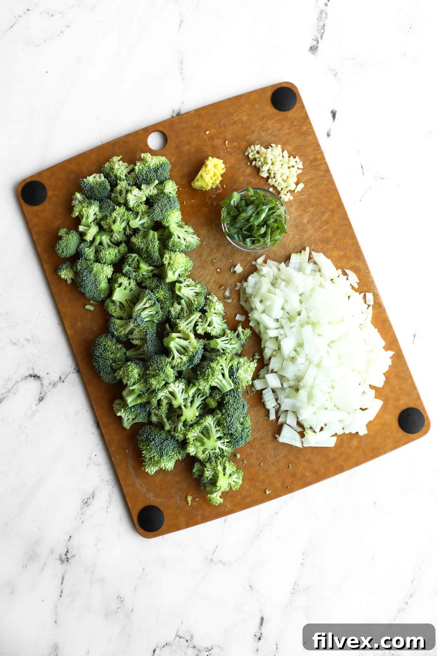 Chopped broccoli, diced onion, sliced green onions, minced garlic, and grated ginger on a brown cutting board.