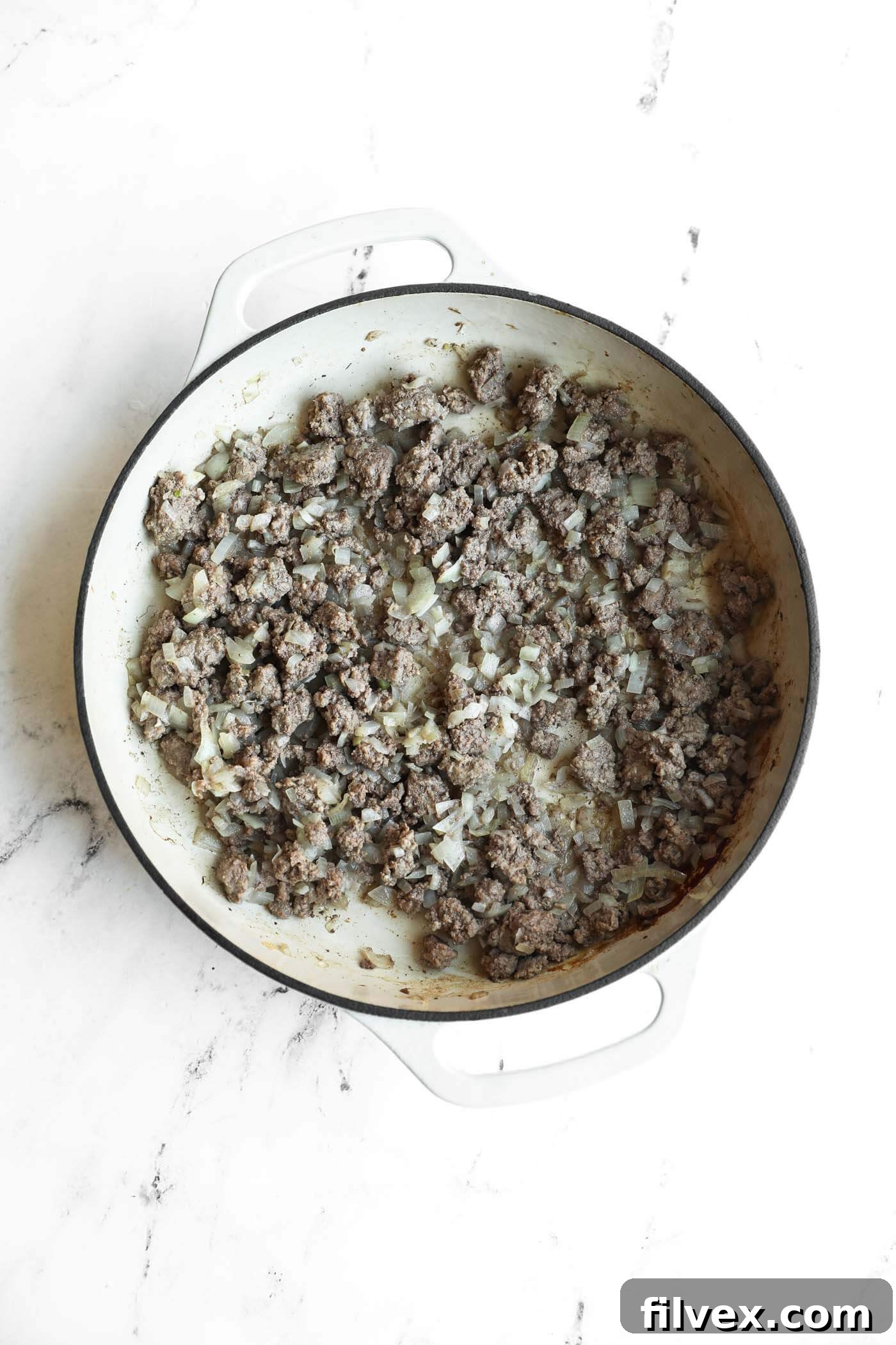 A white pot on a marble surface contains cooked ground beef mixed with chopped onions.
