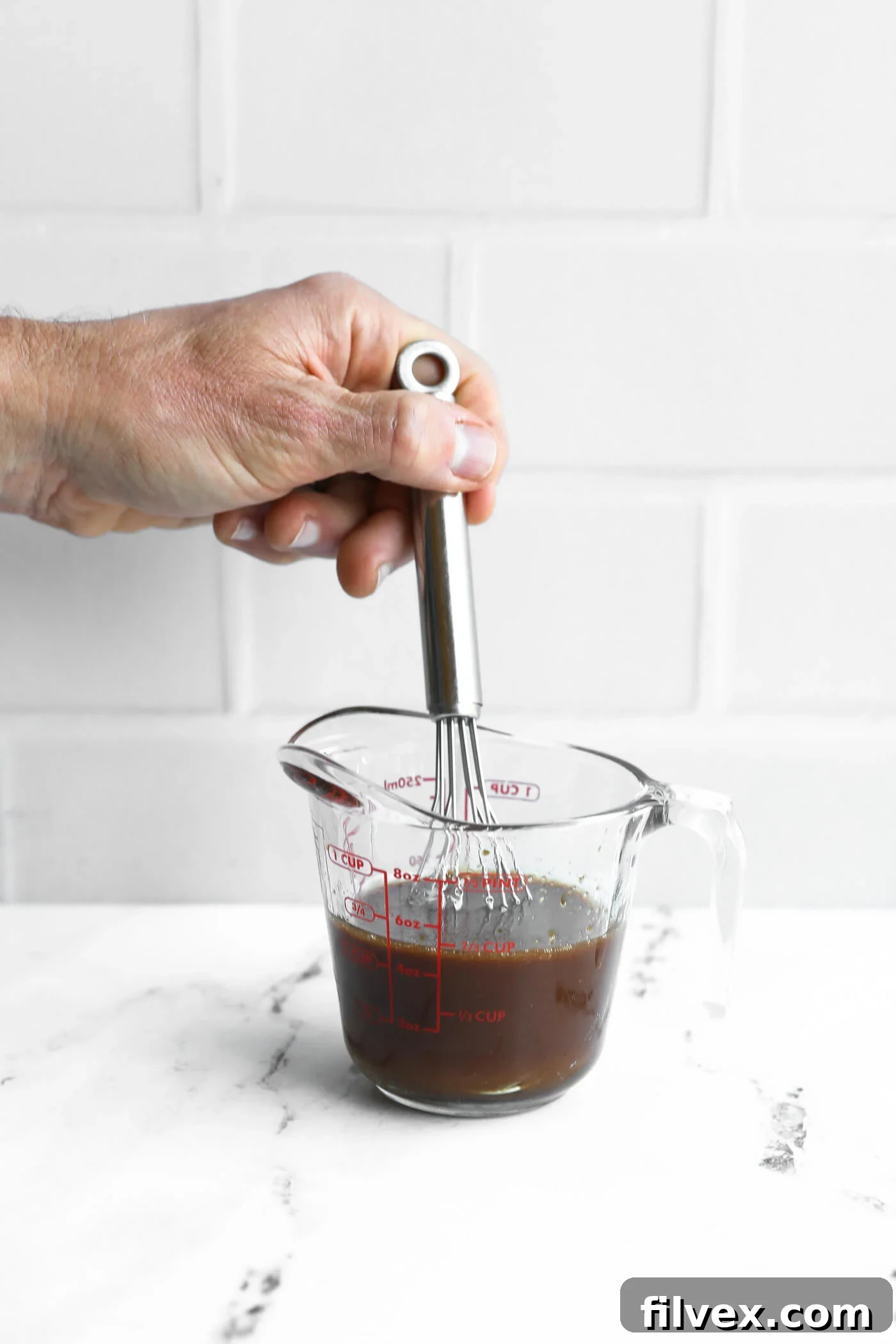 A hand holds a whisk in a glass measuring cup filled with dark liquid on a white countertop with a white tile background.
