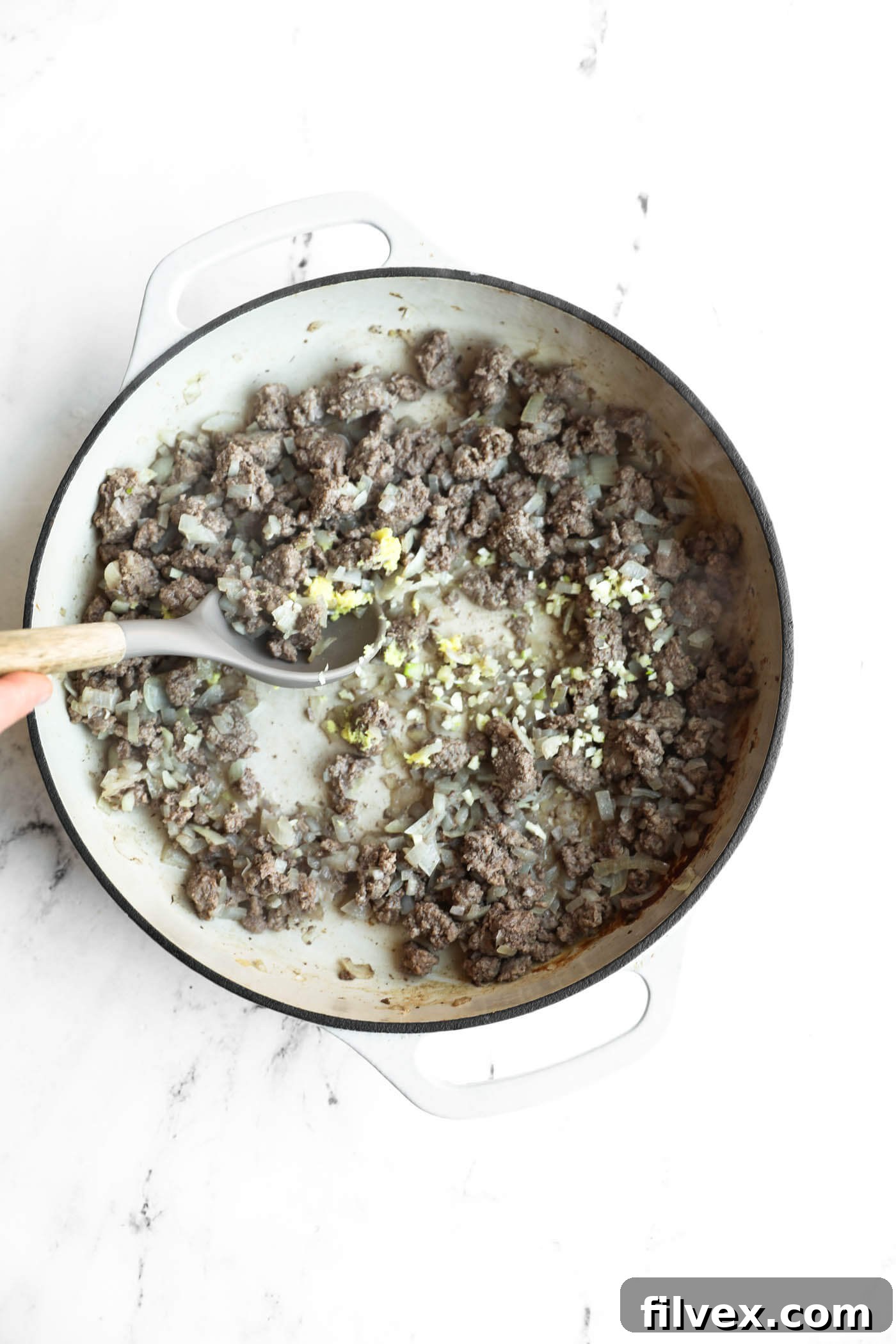 Ground meat, chopped onions, and minced garlic are being sautéed in a white pot on a white surface, with a hand stirring using a wooden spoon.
