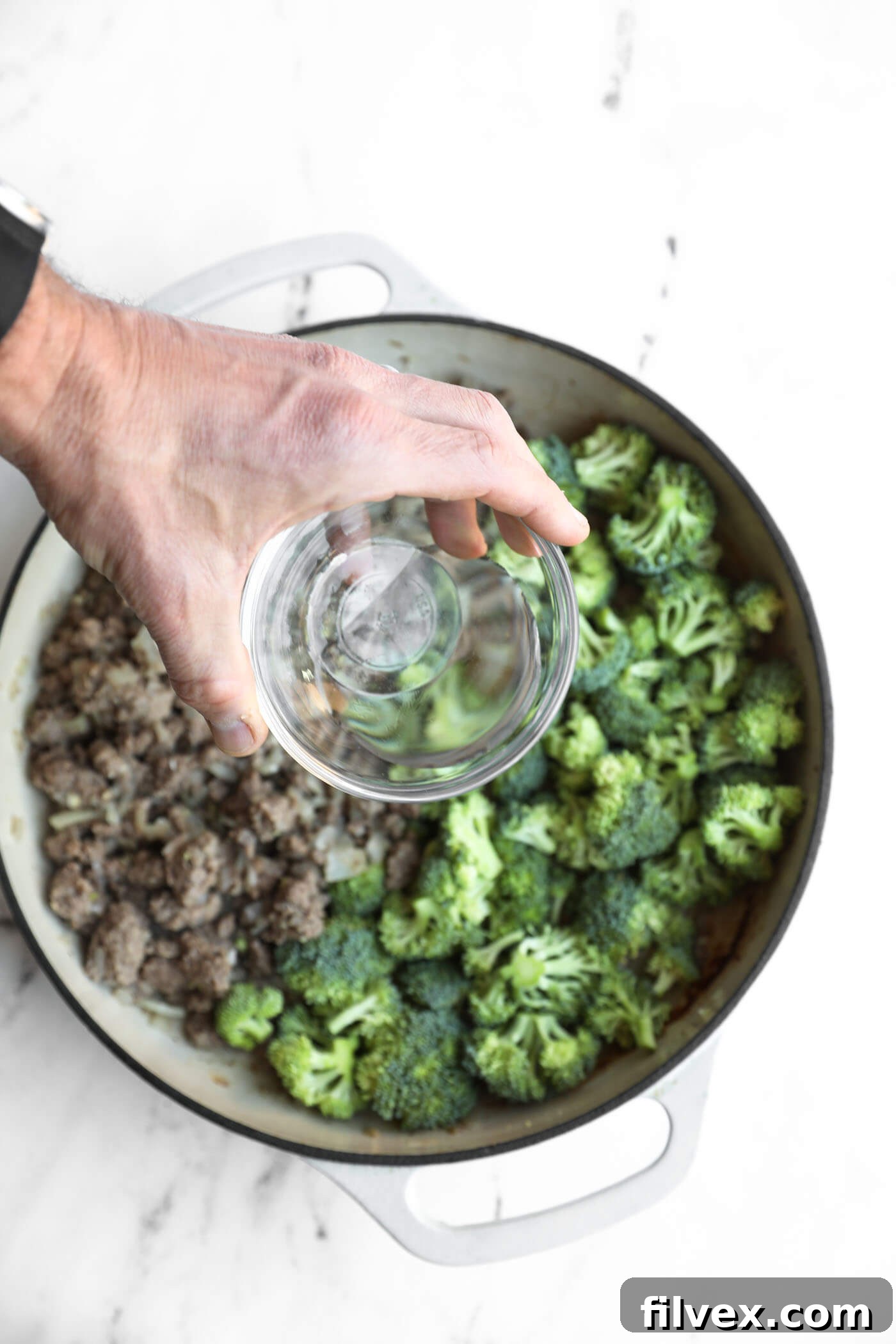 A hand pours water into a pot containing cooked ground meat and fresh broccoli florets.
