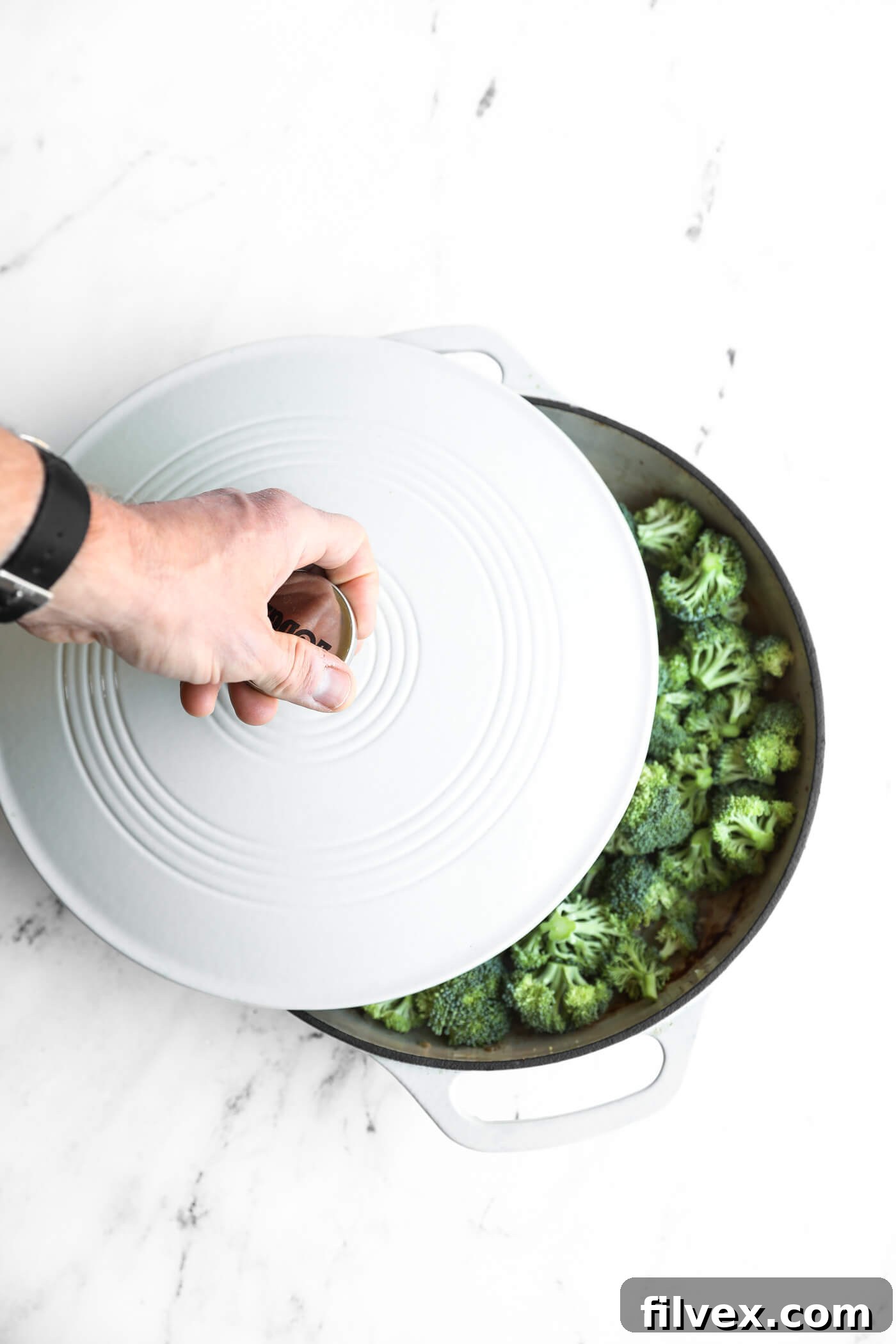 A hand lifts the lid of a white Dutch oven revealing chopped broccoli inside on a white marble surface.