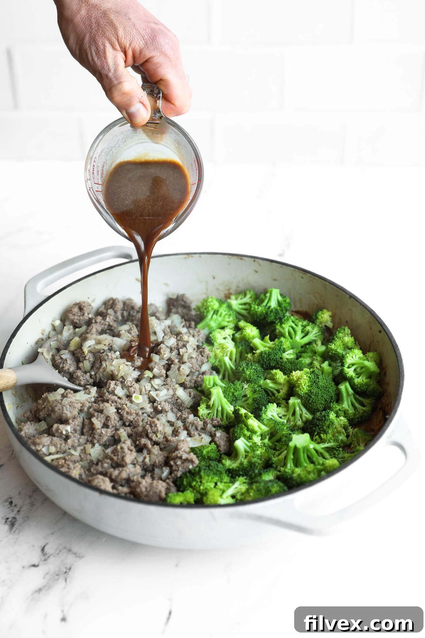 A hand pours brown sauce from a measuring cup into a skillet containing cooked ground meat, onions, and broccoli florets.
