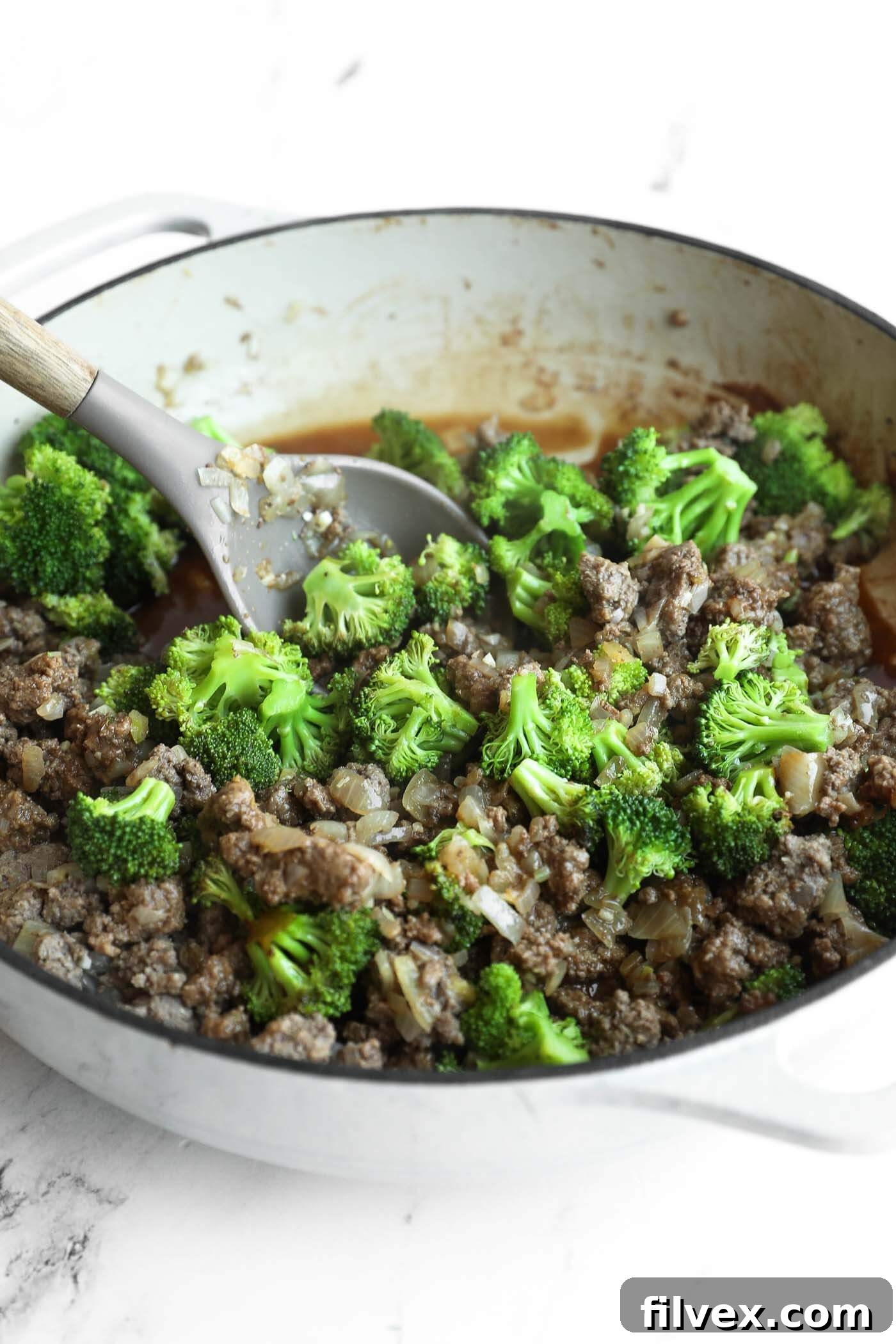 A white pot filled with cooked ground beef, chopped onions, and broccoli florets, with a spoon resting inside.