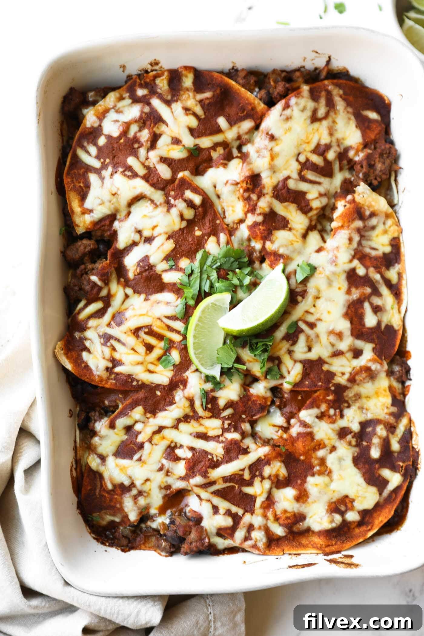 A baked casserole dish filled with cheese-topped enchiladas, garnished with lime wedges and cilantro.