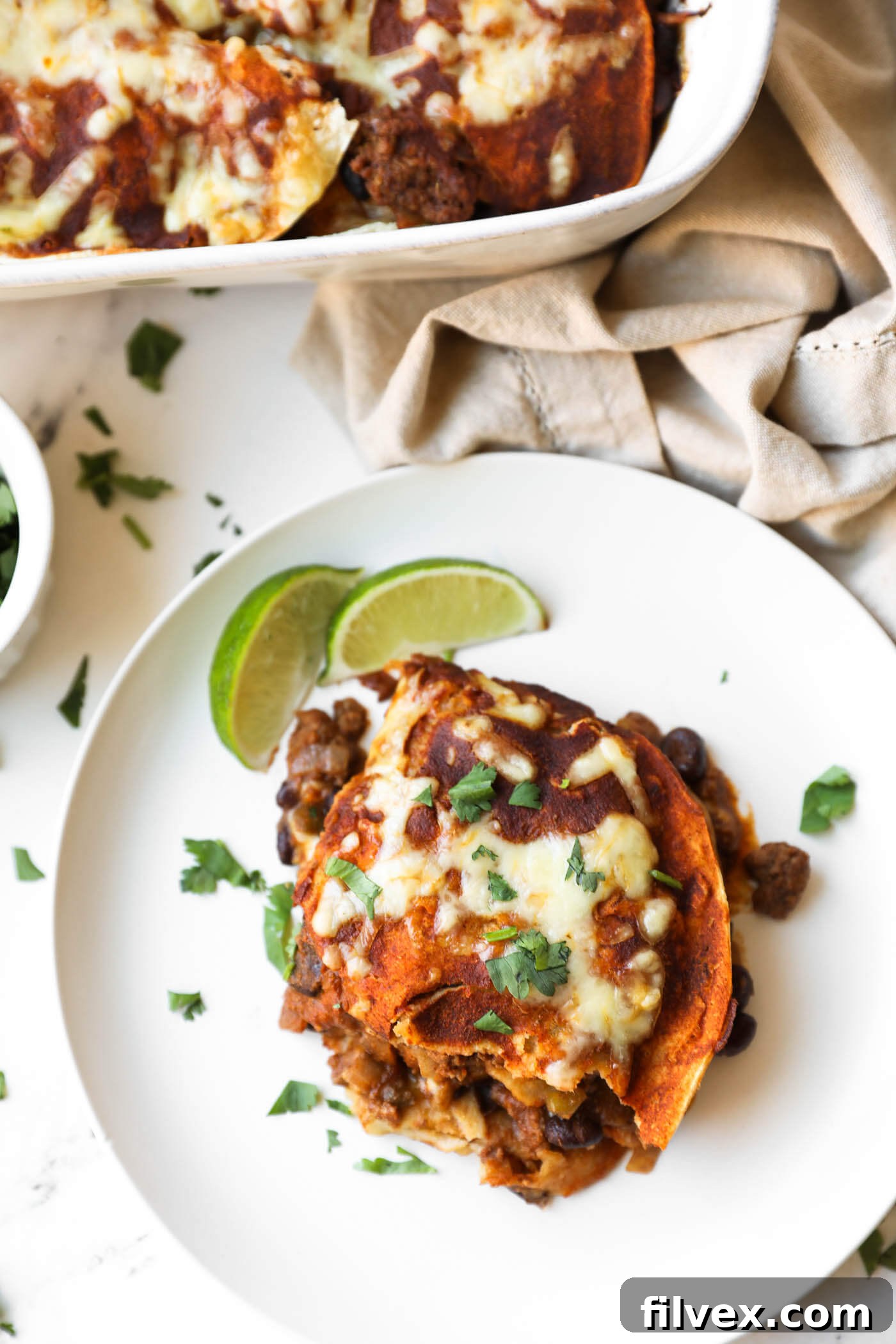 A plate with a serving of cheesy enchiladas topped with chopped cilantro, black beans, and two lime wedges, next to a baking dish with more enchiladas.