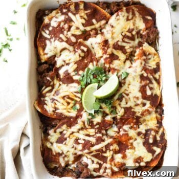 A baked dish of enchiladas topped with melted cheese and garnished with lime wedges and cilantro, placed in a white baking dish on a marble surface.