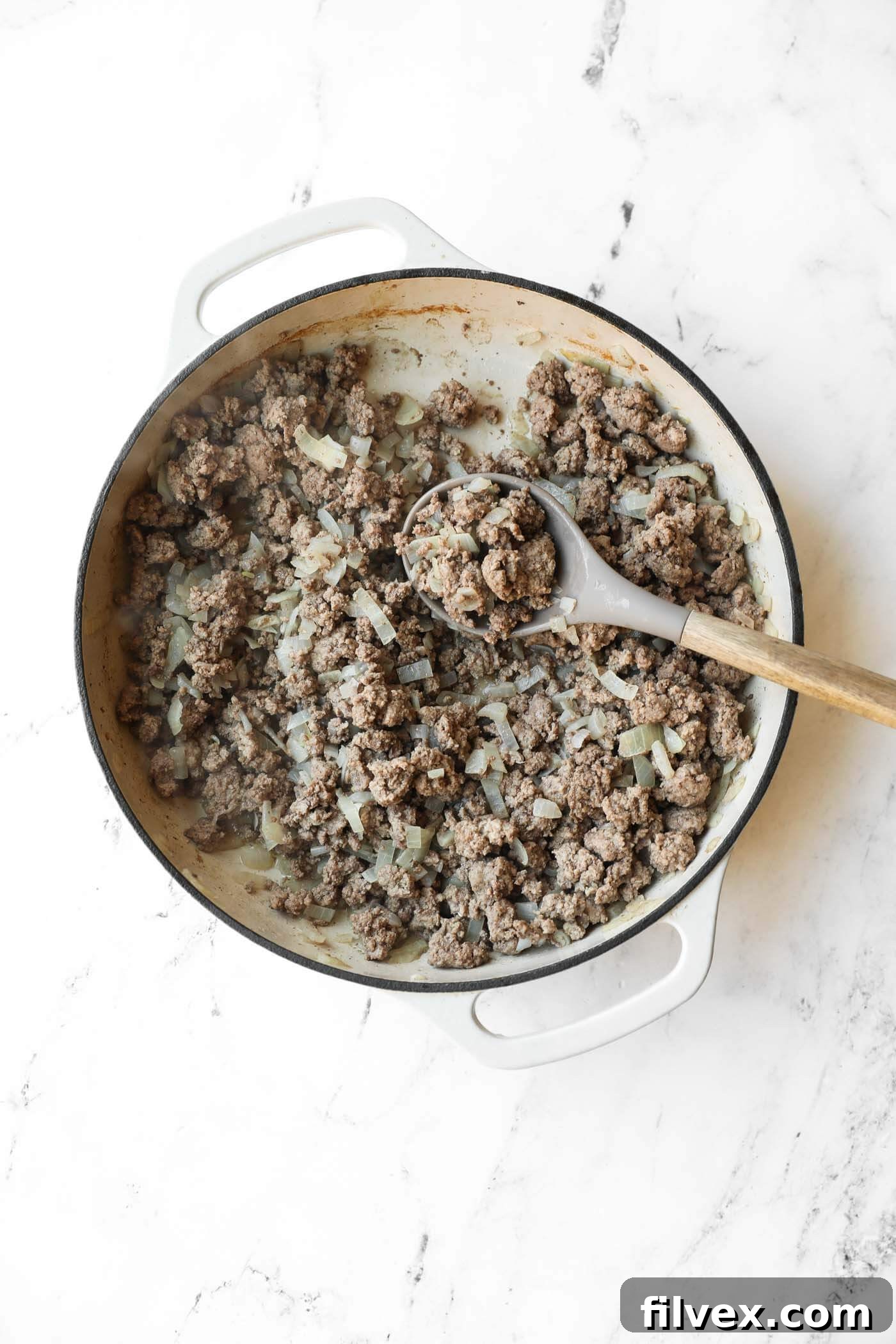Cooked ground beef and chopped onions in a white cast iron skillet, with a wooden spoon resting in the pan, on a white marble surface.