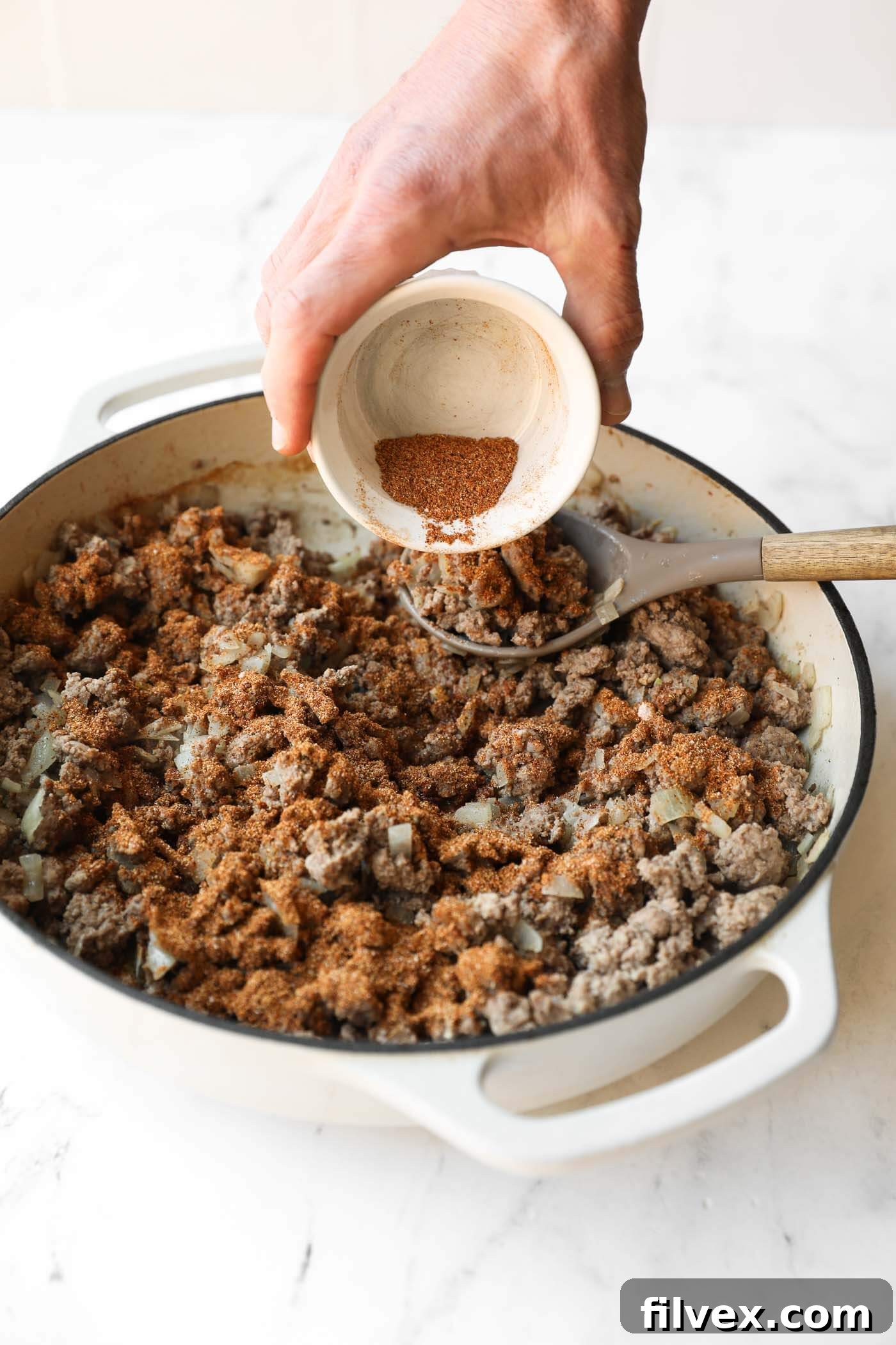 A hand pours spice mix from a small bowl onto cooked ground meat and onions in a white skillet on a marble countertop.