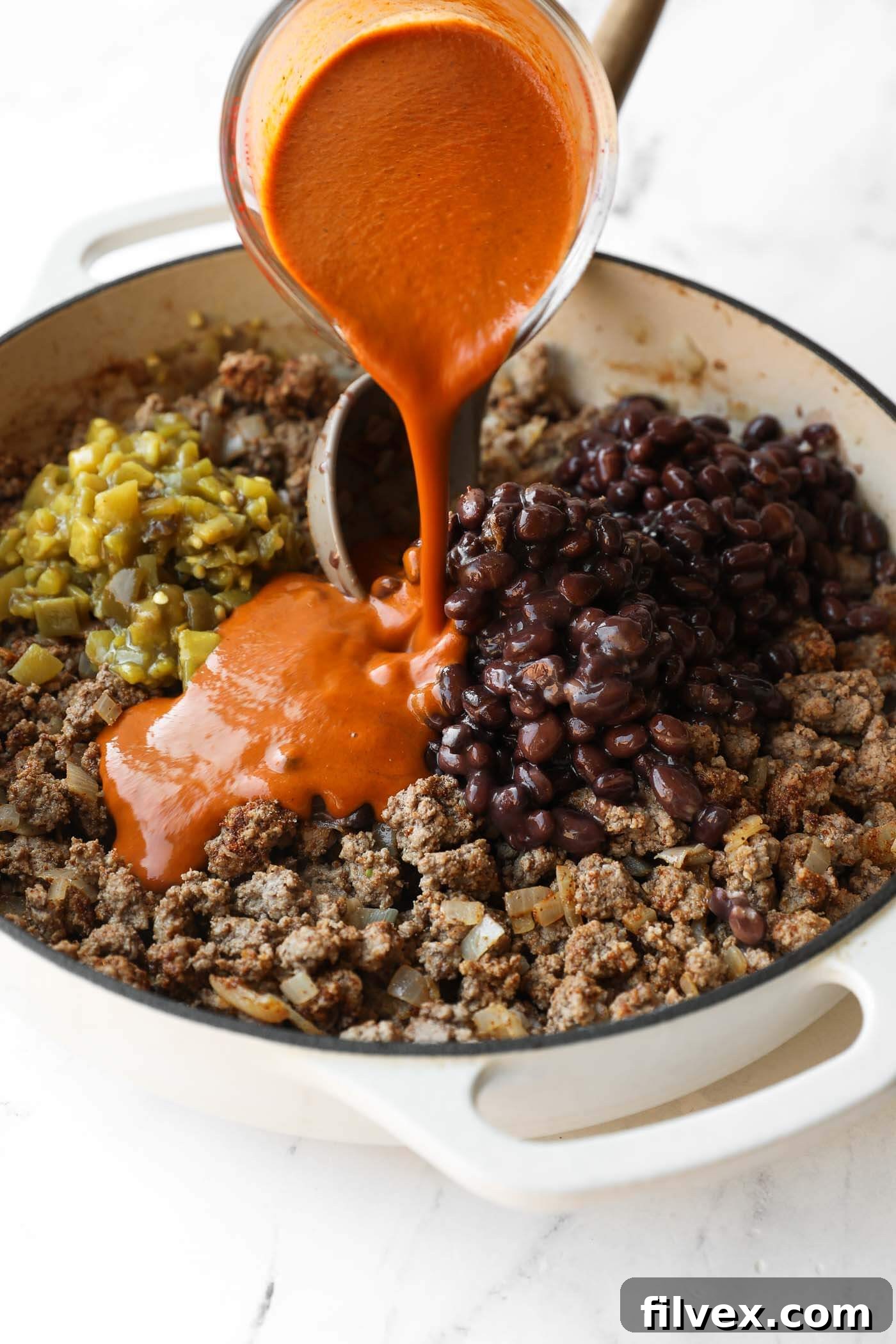 A pot with cooked ground meat, black beans, chopped green chiles, and red sauce being poured in from a measuring cup.