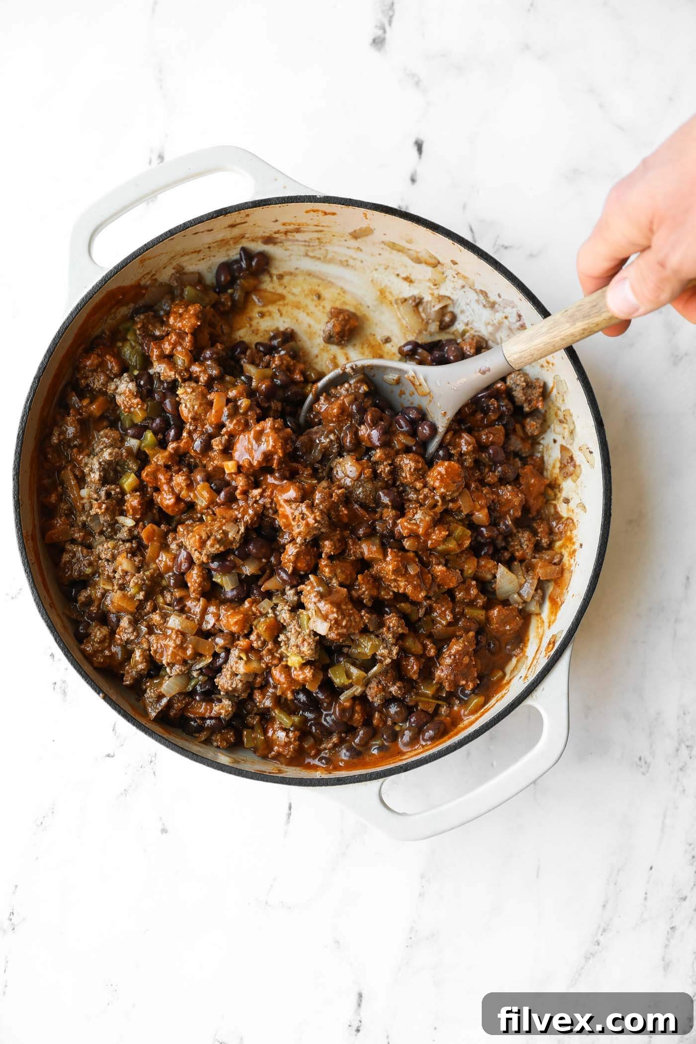 A hand stirs a mixture of ground meat, black beans, and sauce in a white pot on a marble countertop.
