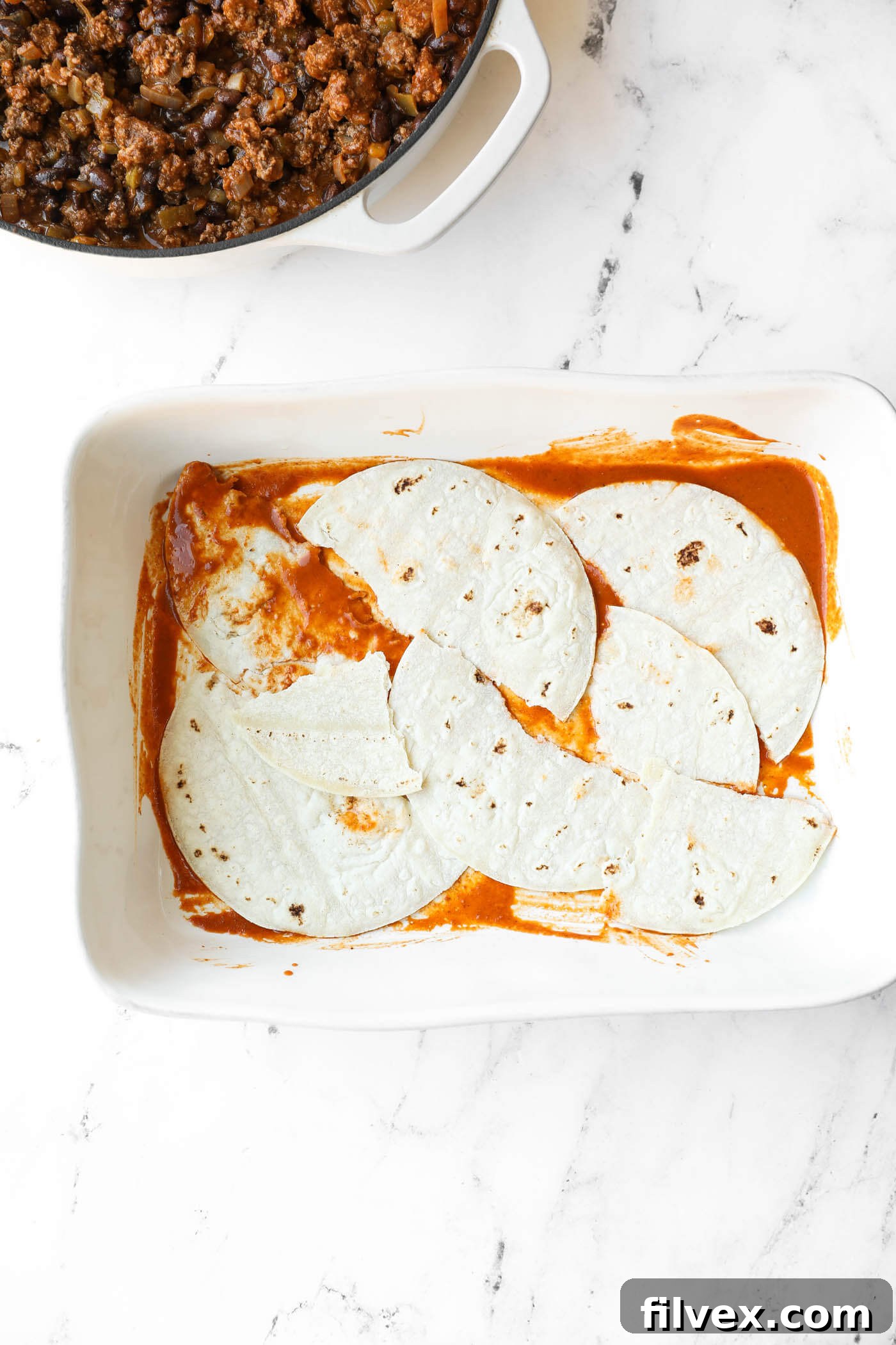 A white baking dish with corn tortillas layered over red sauce, next to a pot of cooked ground meat mixture on a white marble surface.