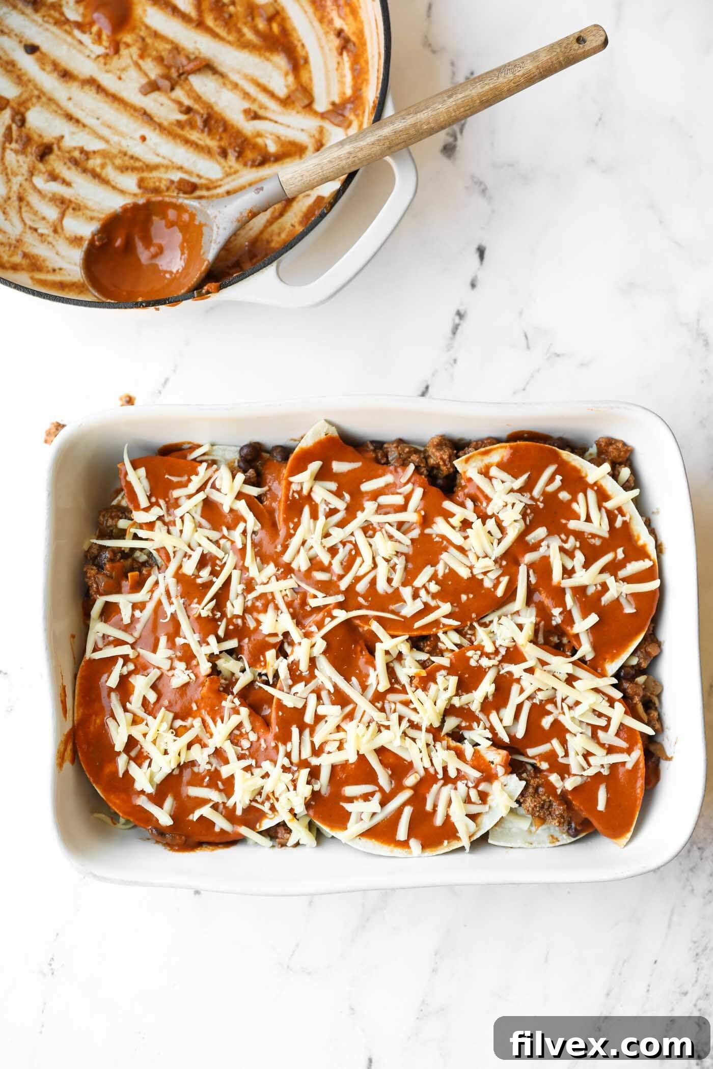 A white baking dish filled with a layered casserole topped with sauce and shredded cheese sits on a marble surface; an empty pot with sauce residue is beside it.